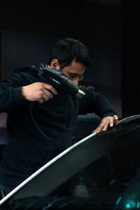 Mechanic carefully applies heat to car windshield with a heat gun in a garage setting.